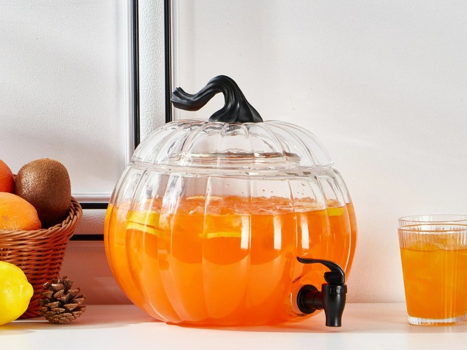 glass pumpkin shaped beverage dispenser filled with orange punch on a table next to a cup of juice