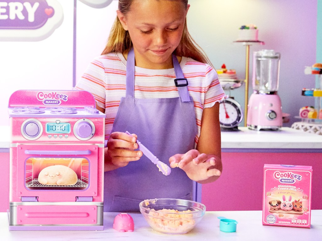 girl putting ingredients into mixing bowl next to a pink Cookeez Makery Oven