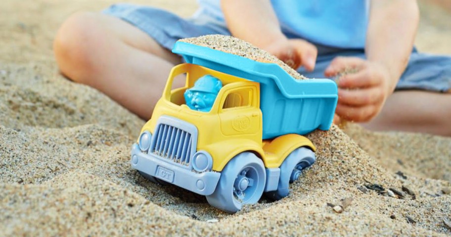child putting sand into back of blue and yellow dump trunk toy