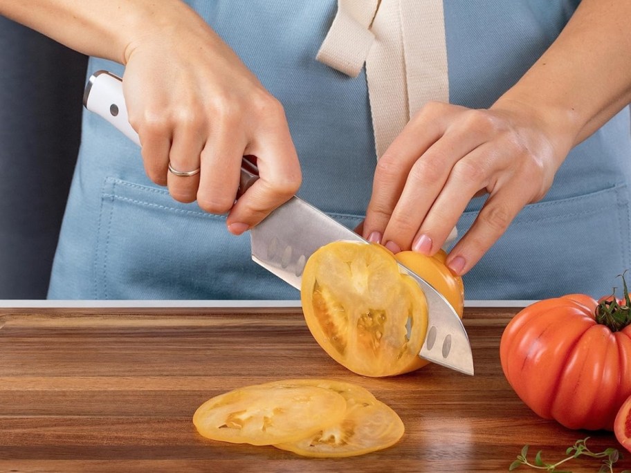 woman using a large chef's knife with a white handle to thinly slice a yellow tomato on a cutting board