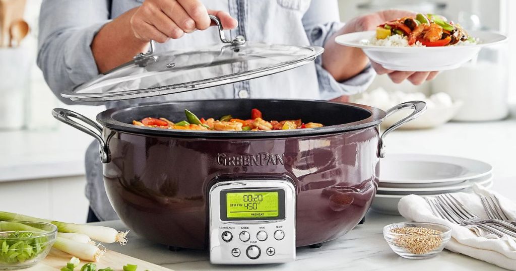 GreenPan Elite 6-Quart Electric Multi-Pot with Steamer & Tool shown in red being used by a woman to cook a meal