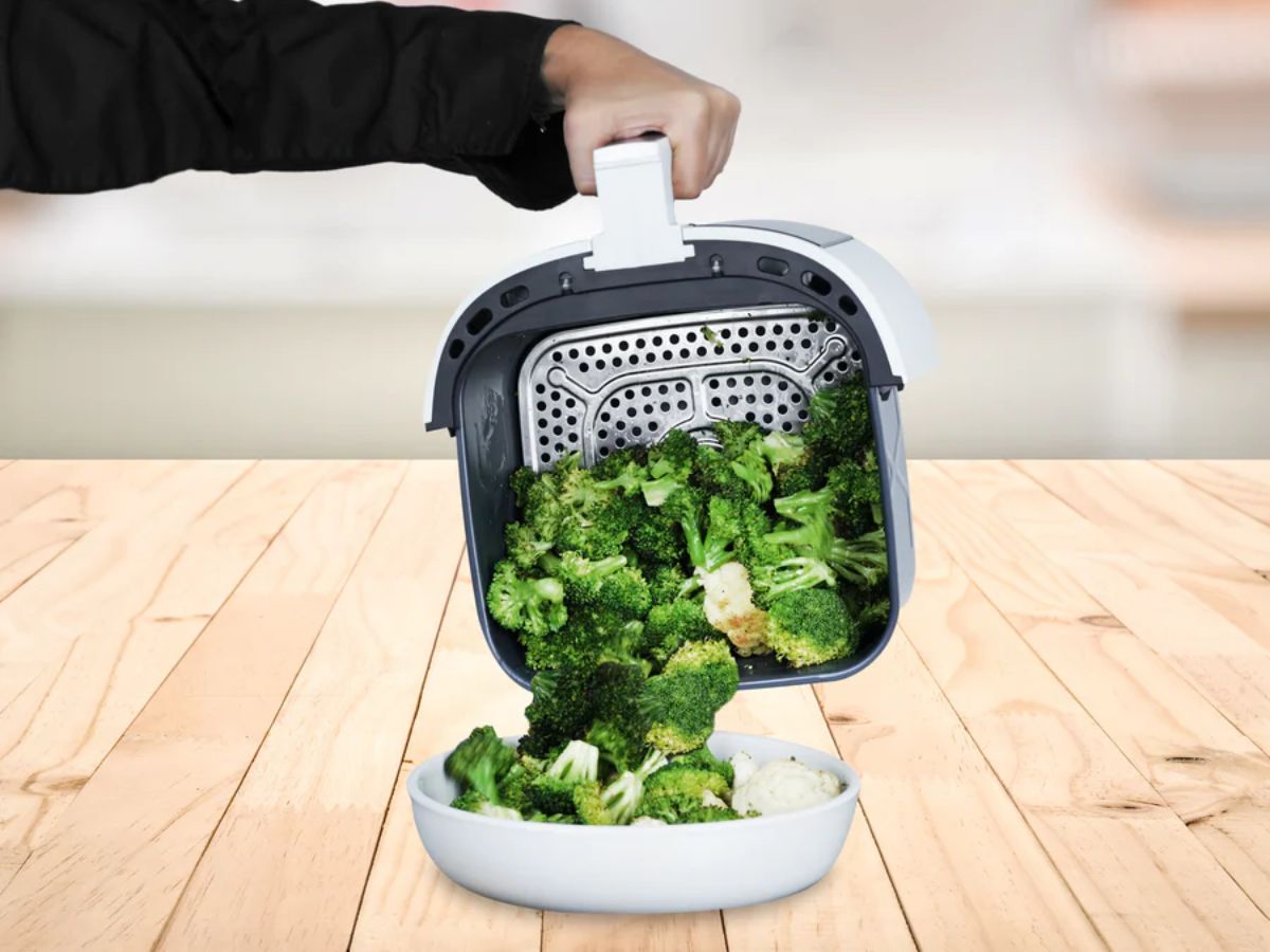 a womand hand holding an airfryer basket and pouring broccoli florets out of the basket into a white bowl 