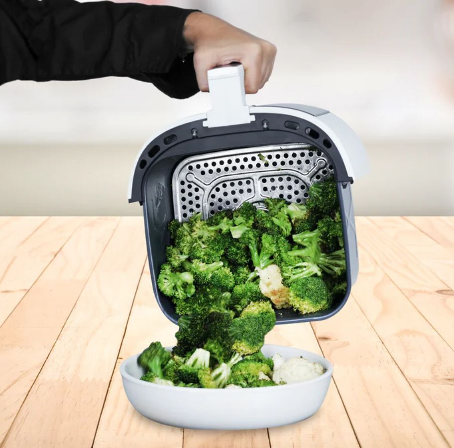 a womand hand holding an airfryer basket and pouring broccoli florets out of the basket into a white bowl 