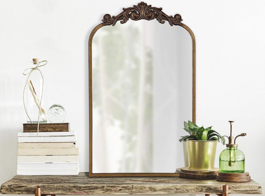 an arched mirror with gold trim on a rustic console table next to plants and a stack of books