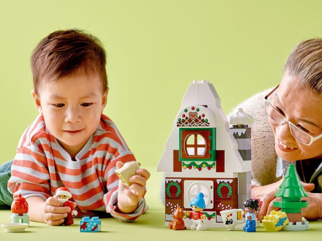 toddler boy playing with a LEGO DUPLO Santa's Gingerbread House while a woman wearing glasses watches from the side