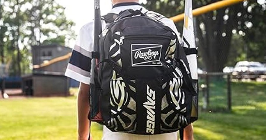 A person standing outdoors with a black backpack that features the logos "Rawlings" and "SAVAGE," holding baseball bats.