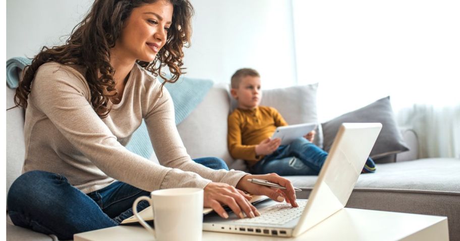 Woman sitting on a couch using a laptop with a little boy in the background using a tablet