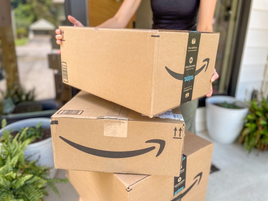 woman lifting an amazon prime box off a stack of other boxes from in front of the house