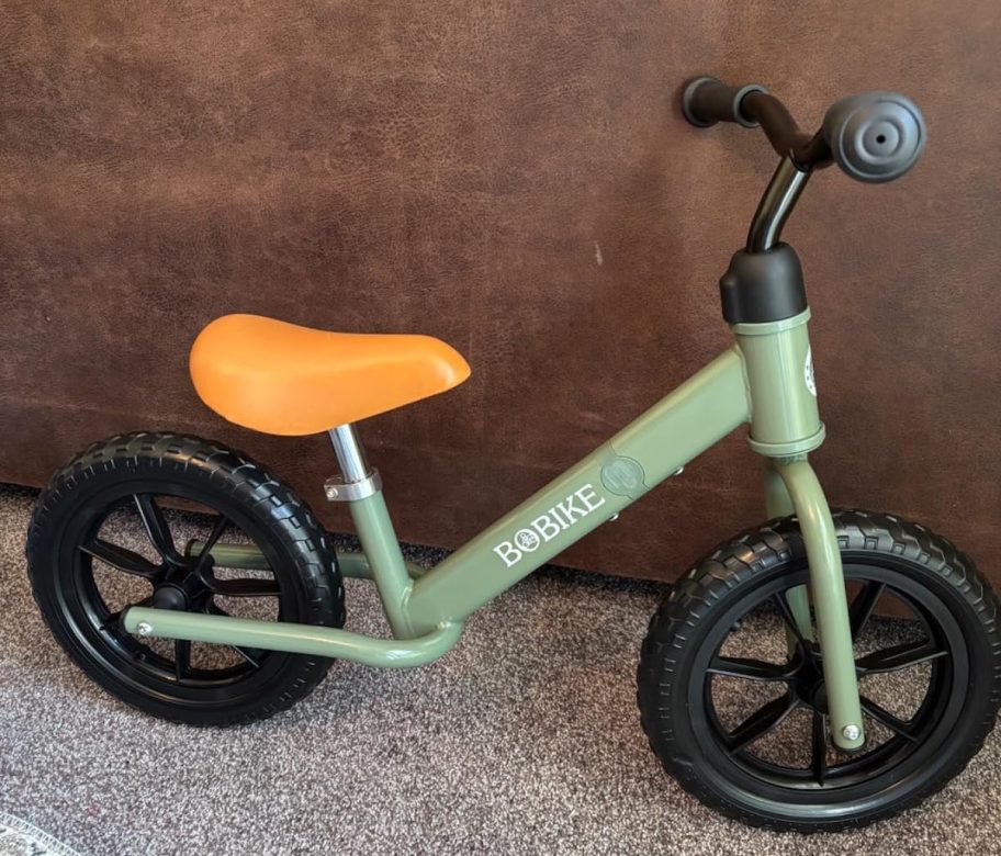 Green toddler, balance, bike, sitting on carpet, leaning on Brown wall