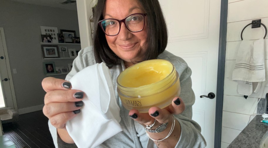 smiling woman wearing a robe holding an open jar of colleen rothschild cleansing balm