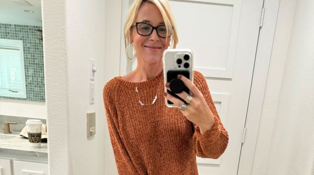 Woman taking selfie in hallway wearing name, necklace, and brown sweater 