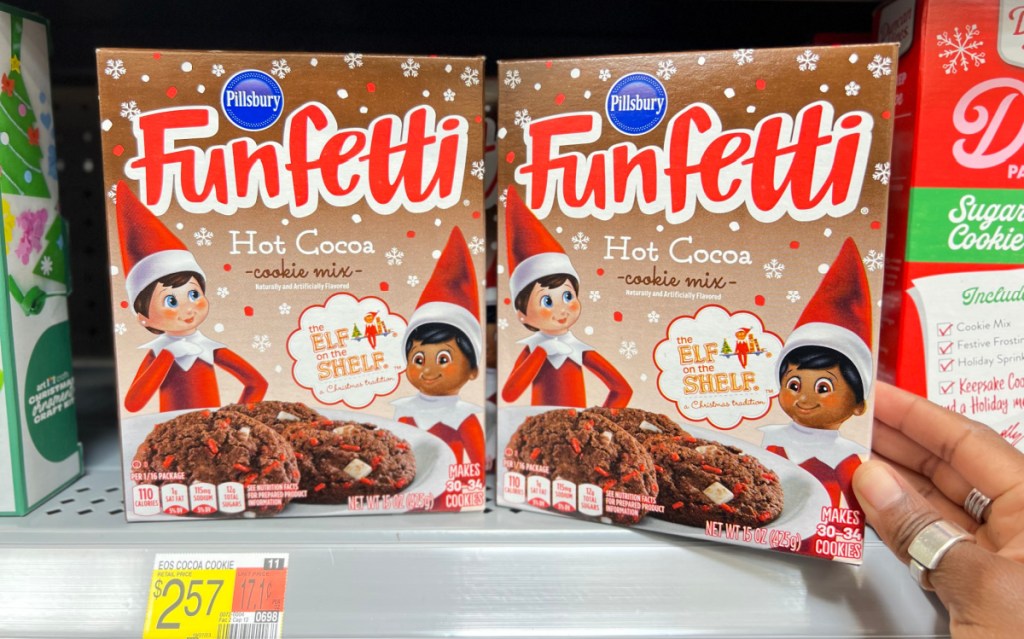 two boxes of pillsbury funfetti elf on the shelf hot cocoa cookie mixes on a walmart shelf being touched by a hand