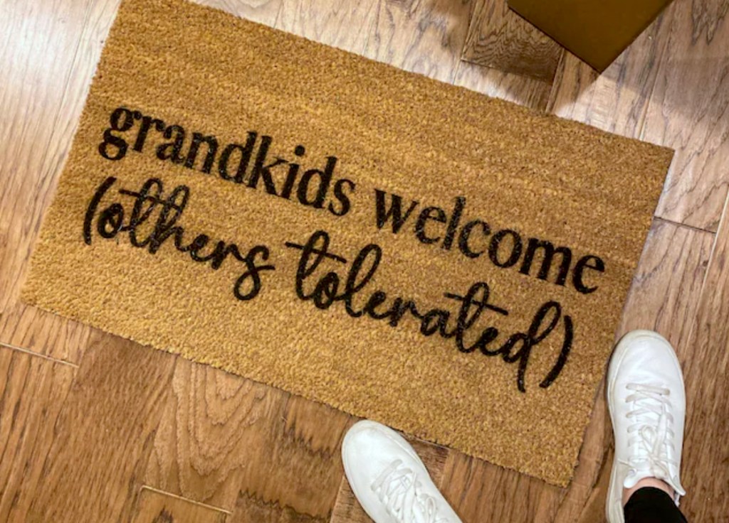 two feet standing on wood floor next to doormat that says grandkids welcome others tolerated