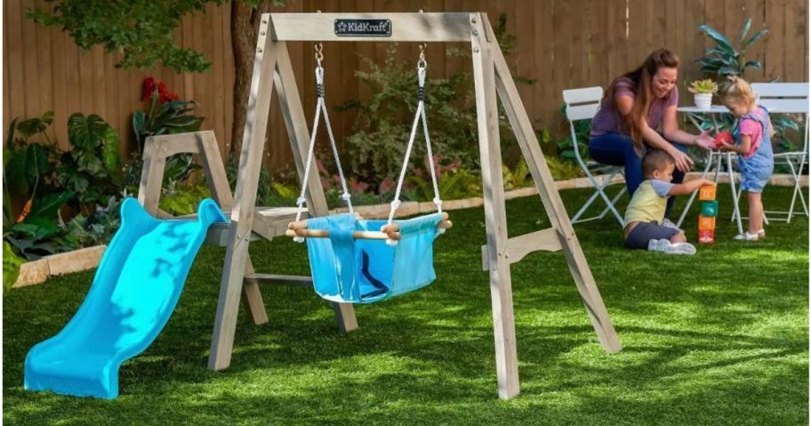 KidKraft My First Wooden Swing Set on lawn with mother and daughter in the background
