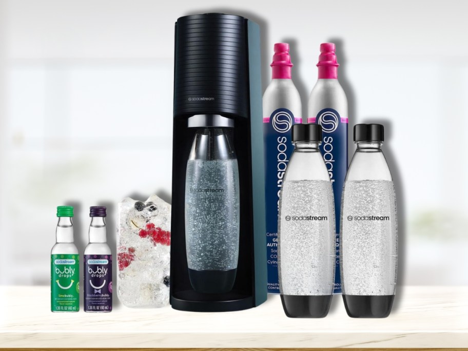 black Sodastream machine with 2 CO2 containers, 2 reusable bottles, a glass of soda and 2 Bubly flavor bottles on a counter