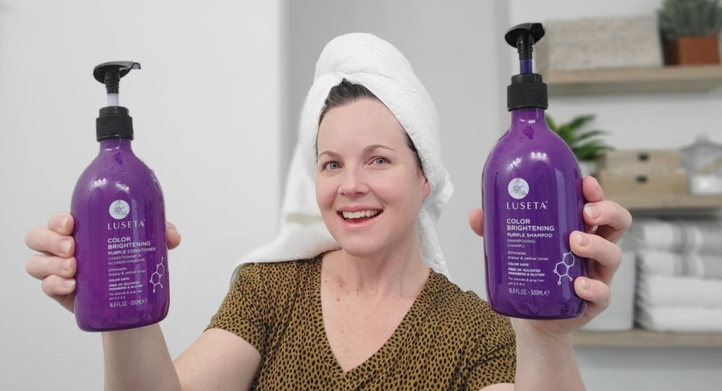 woman holding up purple shampoo and conditioner while in the bathroom with a shower towel on her head