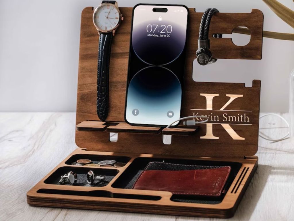 قاعدة توصيل خشبية مع نقش مخصص وعليها ساعة رجالية وهاتف ومحفظة ومجوهرات