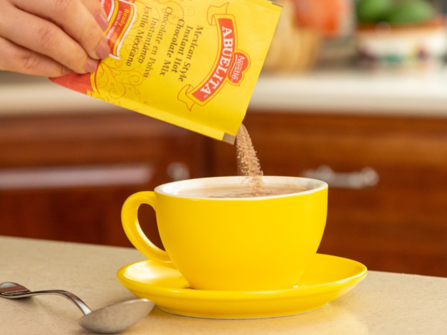 hand pouring Abuelita hot chocolate mix into a yellow mug sat atop a saucer