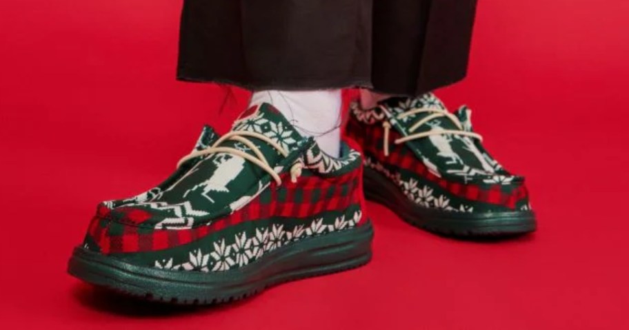 man's legs and feet wearing a pair of red, green, and white ugly sweater style HEYDUDE slip on sneakers - the background of the image is red