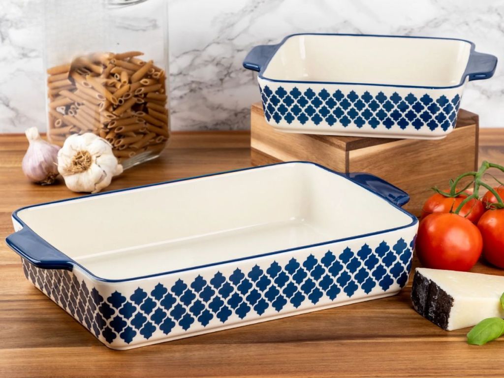 two blue and white casserole baking dishes sitting on counter with tomatoes, cheese, pasta and garlic around them
