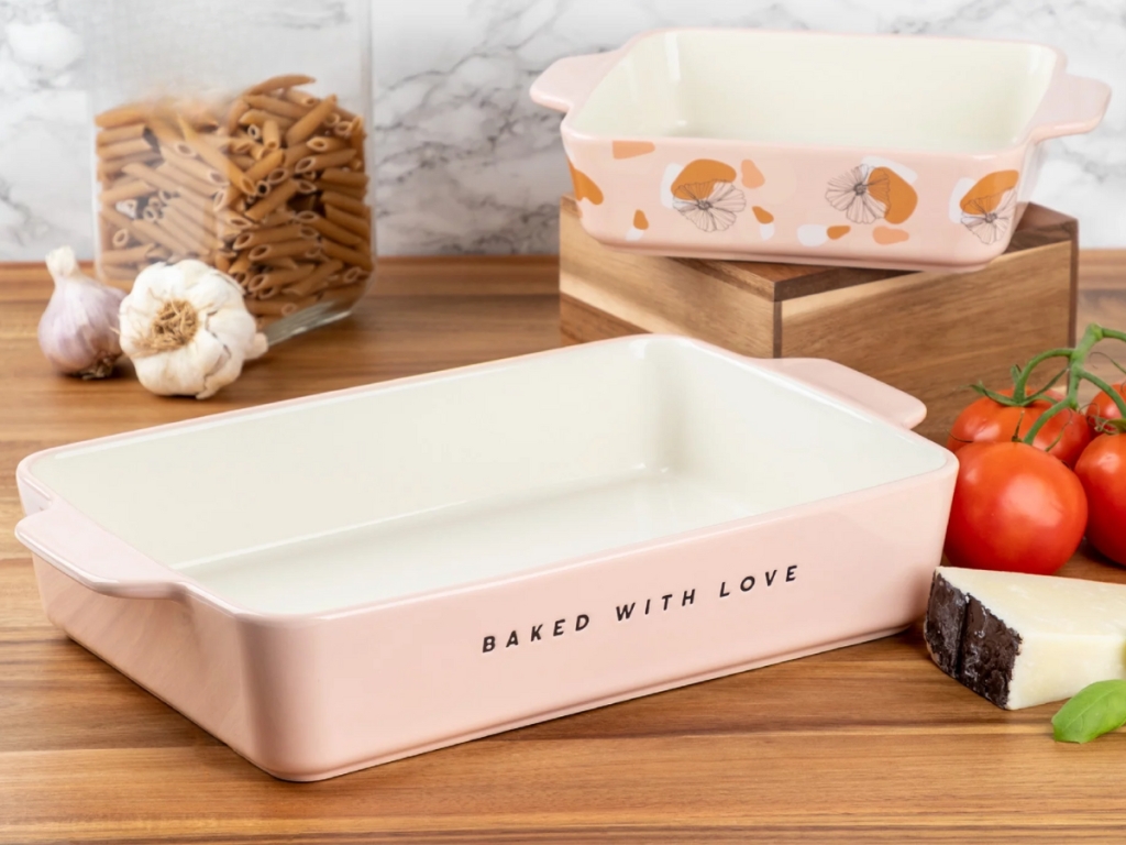 two pink casserole baking dishes sitting on counter with tomatoes, cheese, pasta and garlic around them