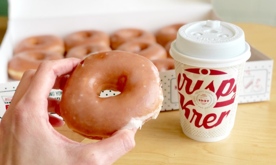 hand holding a Krispy Kreme original glazed donut with a cup of coffee and box of donuts behind him