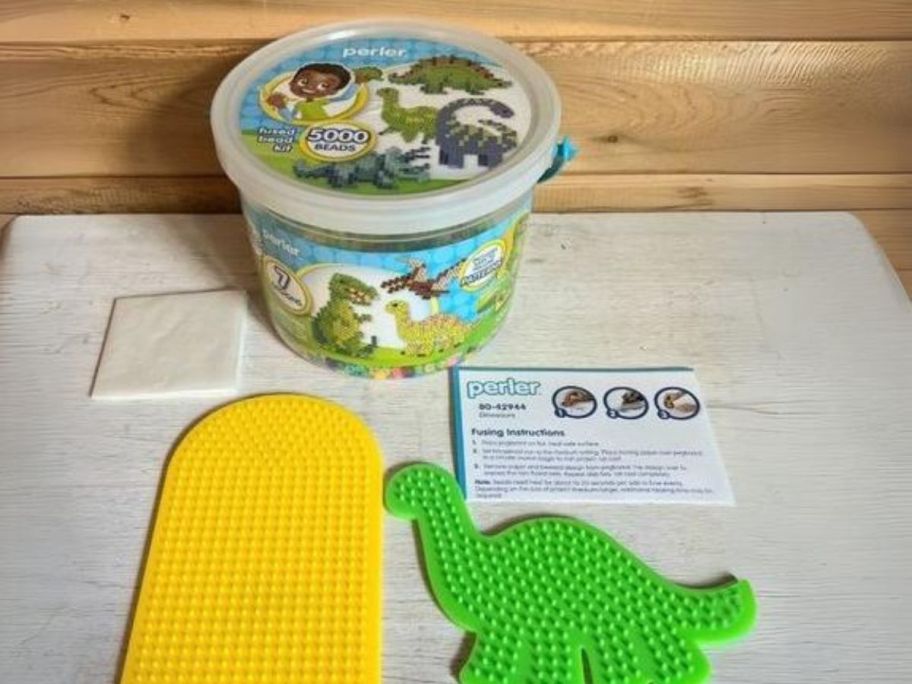 Perler Dinosaur Craft 5,004-Piece Bead Bucket Activity Kit with pegboards, ironing sheet, and instructions sitting in front of it on table