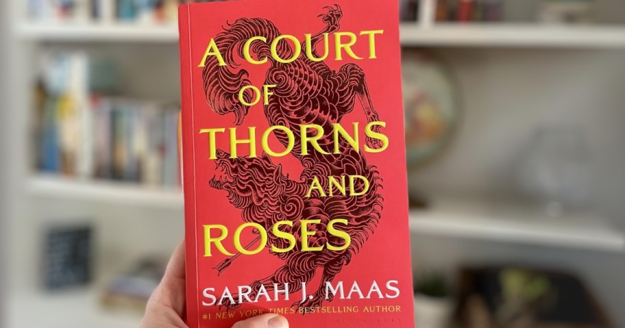 hand holding up the book "A Court of Thorns and Roses" in front of a bookshelf