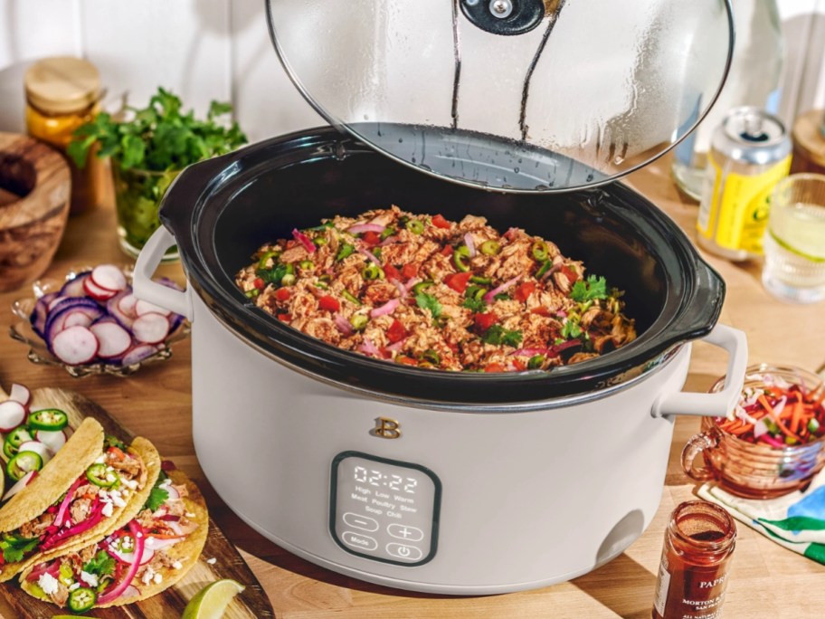 taupe color large slow cooker with food cooking inside and food on plates around it
