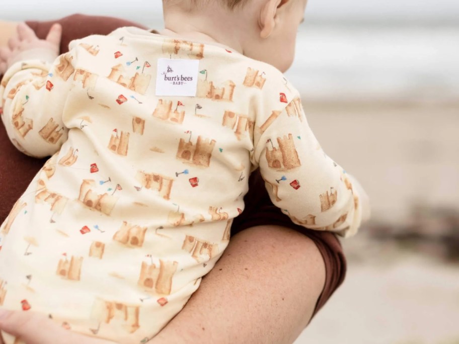 mom holding a baby on the beach, baby is wearing a tan pajama onesie with sand castles on it