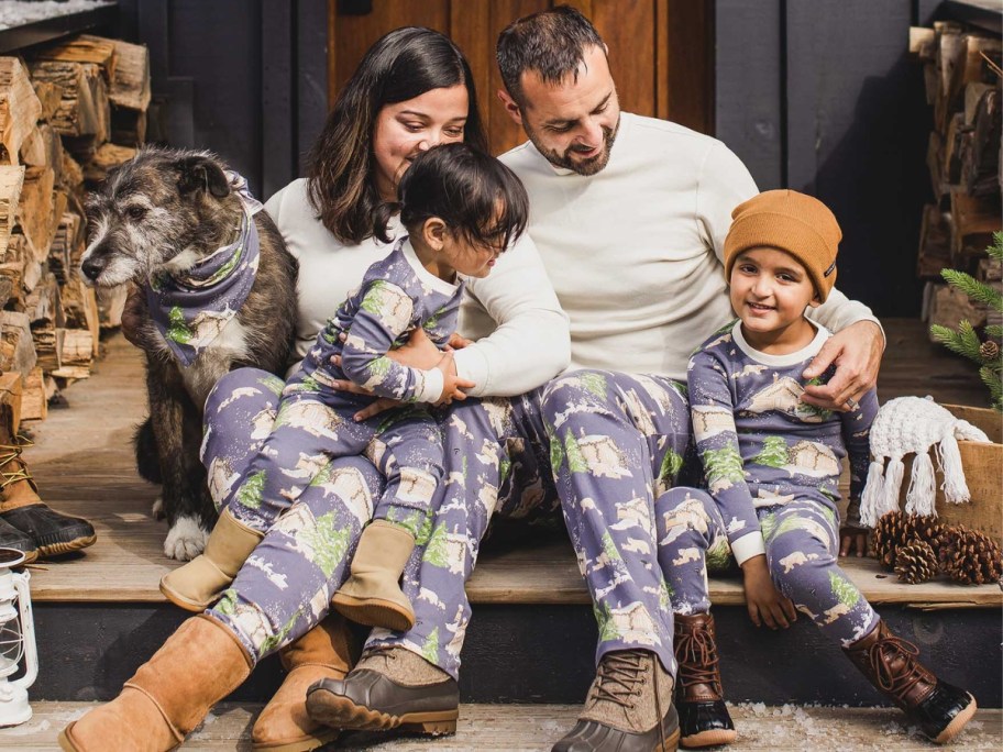 family sitting outside on front steps of a cabin with their dog, all wearing matching blue, green, and white holiday pajamas