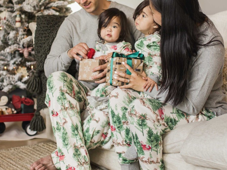 mom, dad, and baby sitting on a couch wearing matching Christmas pajamas, baby is holding a present