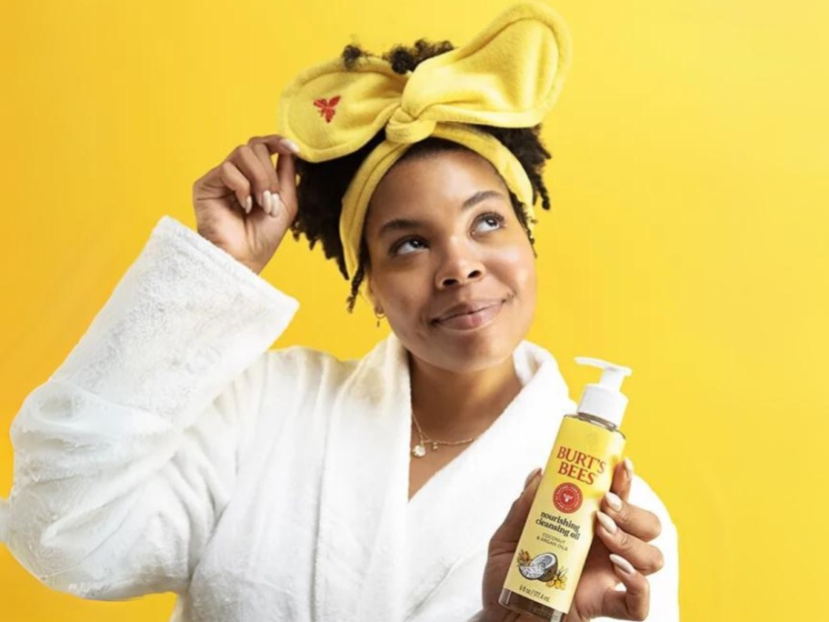 woman wearing a yellow headband and white robe, holding a bottle of Burt's Bees cleansing oil