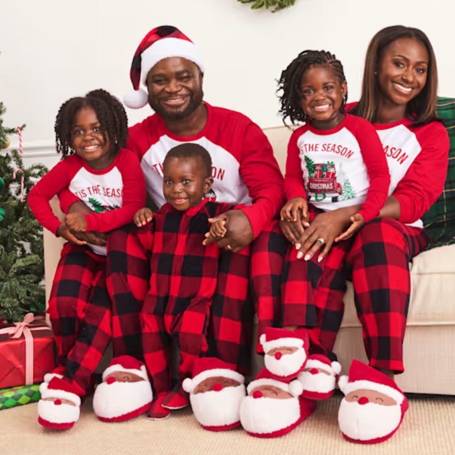 family with mom, dad, 2 little girls and baby boy wearing matching red and black plaid Christmas pajamas that say 