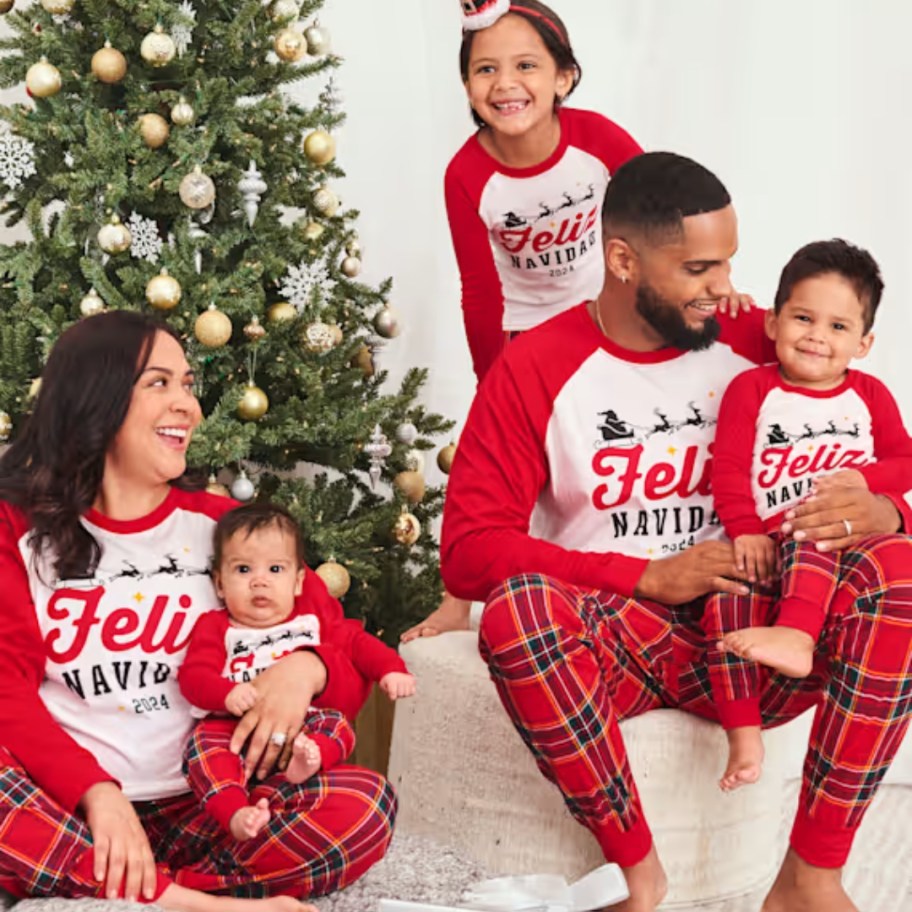 family by a Christmas tree wearing matching red/black plaid and white long sleeve Christmas pajamas that say 