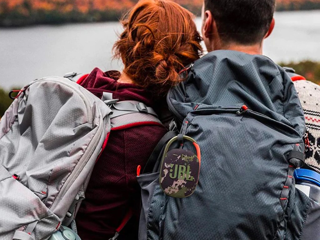 two people looking out at water wearing backpacks with camo jbl speaker hanging on backpack