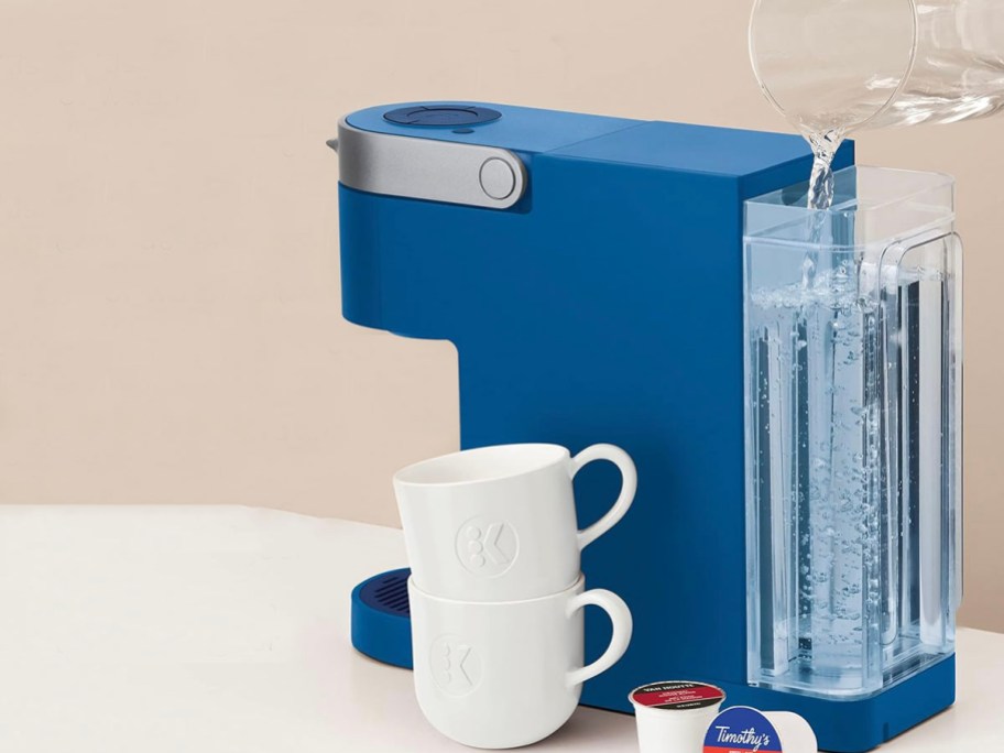 blue keurig slim coffee maker sitting on table with someone pouring water into the reservoir 