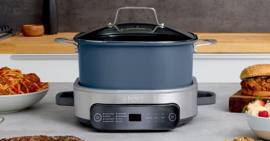 a blue ninja possible cooker on a kitchen counter surrounded by various dishes of food