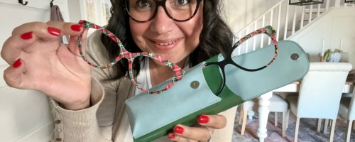 smiling woman wearing glasses and holding up various pair eyewear toppers and a case