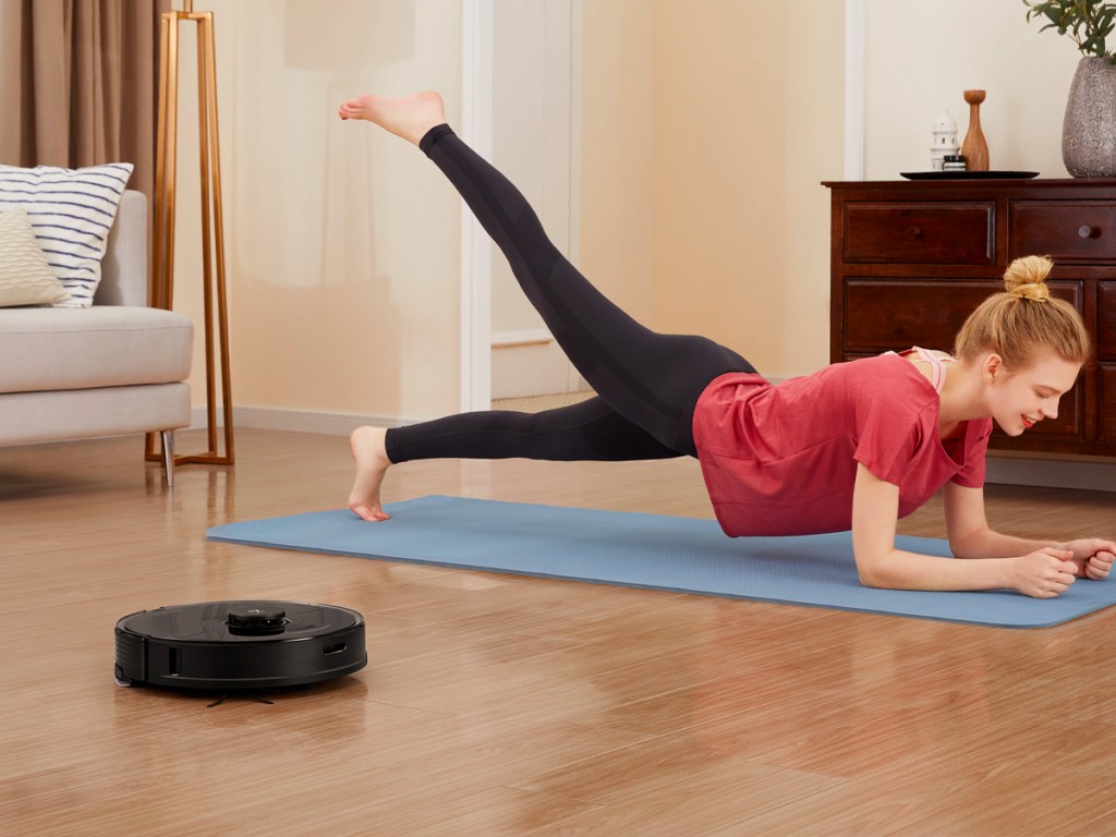 black robot vacuum on floor with woman doing yoga in the background