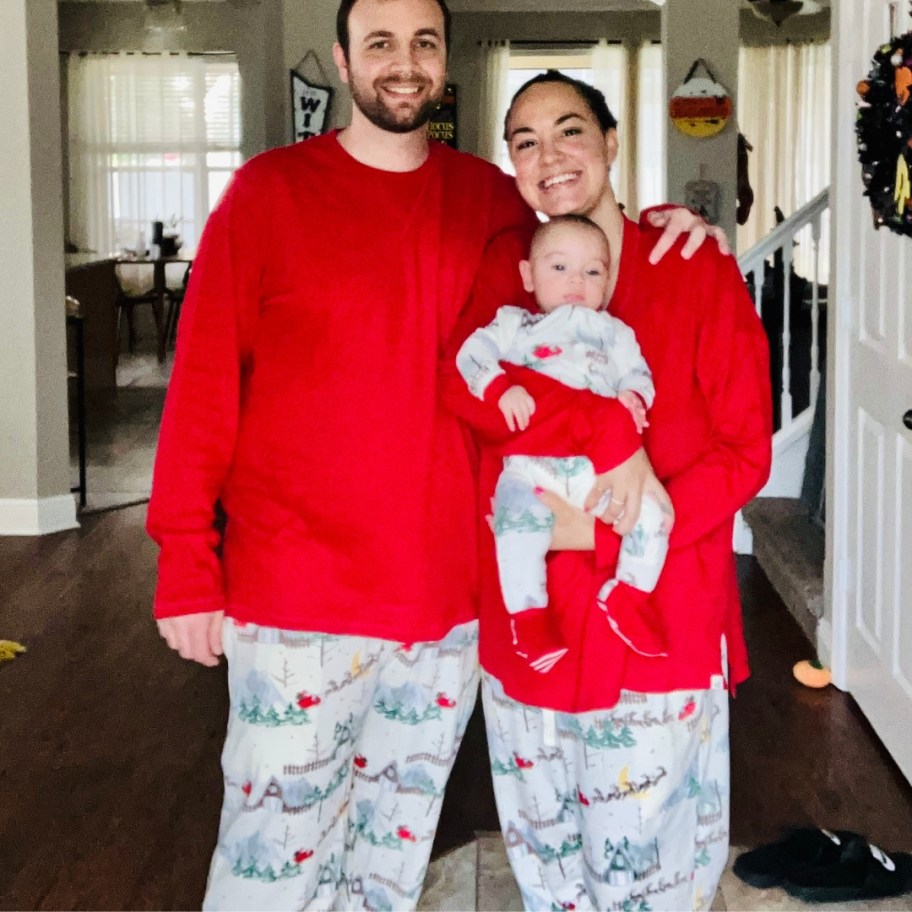 dad, mom, and baby (mom holding the baby) standing wearing matching red, white, and green holiday print pajamas