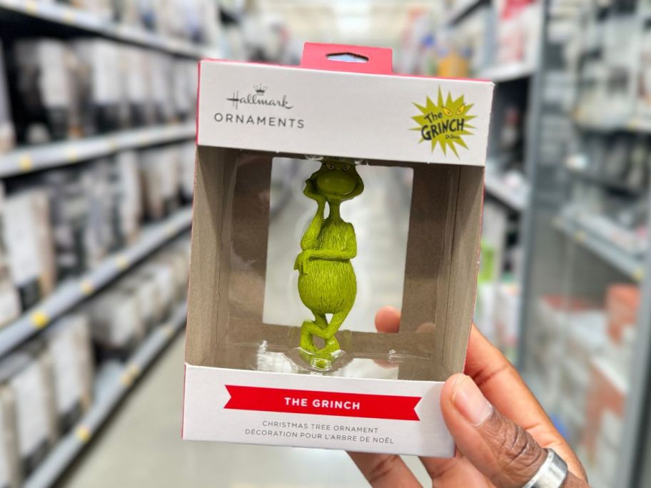 a womans hand holding a boxed grinch ornament