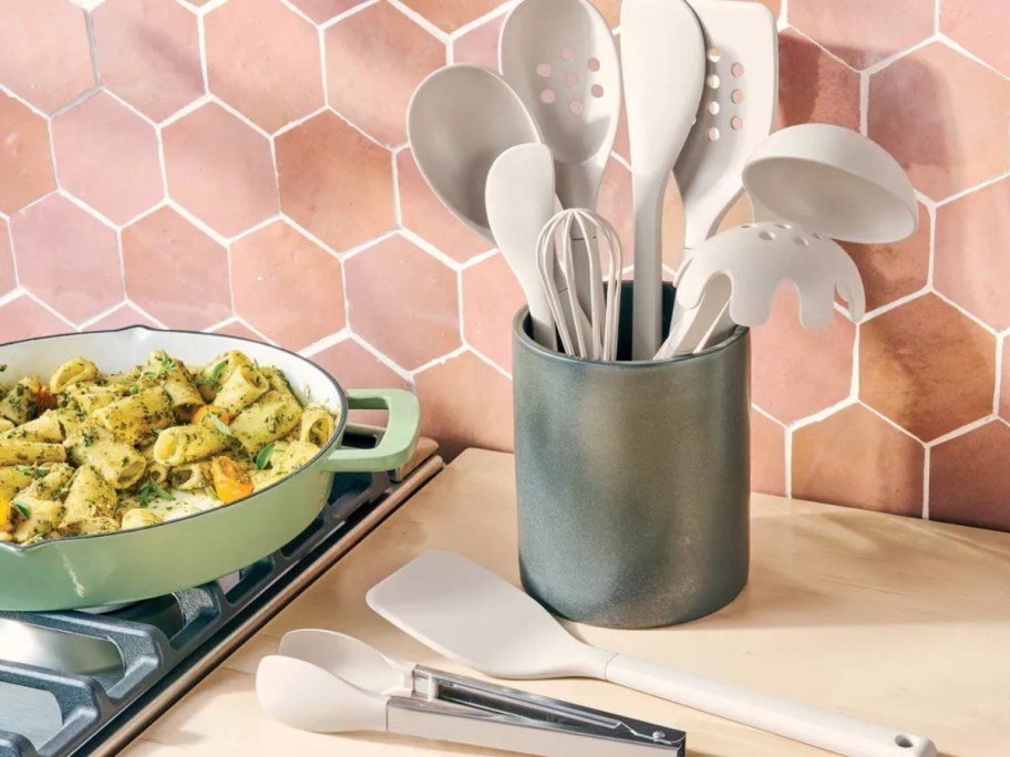 grey kitchen utensils in a green holder with some on the counter next to a stove with food cooking