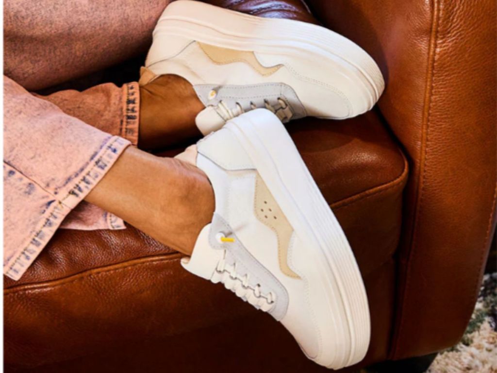 woman's legs and feet hanging off a brown leather sofa, wearing a pair of HEYDUDE Women's Hudson Lift Sport sneakers in white with tan and light blue accents