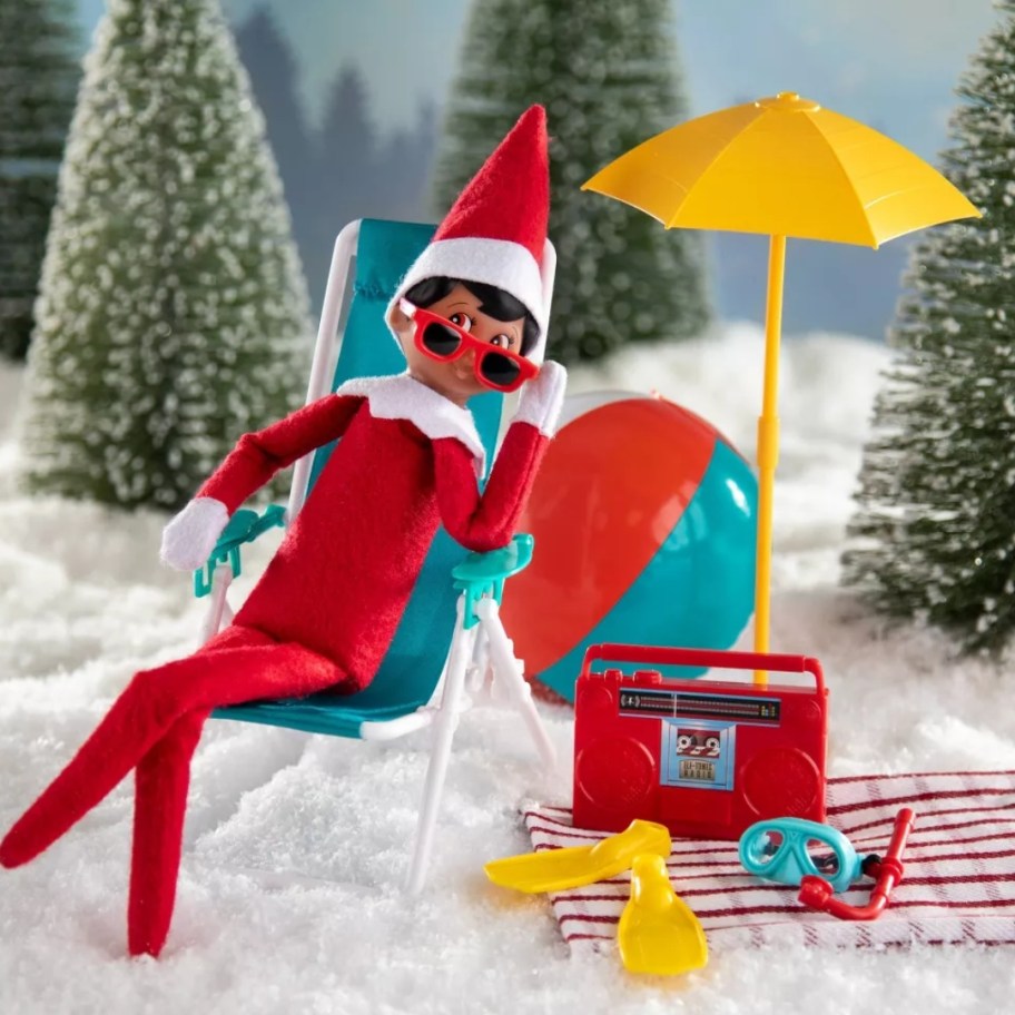 boy Elf on The Shelf doll sitting in a doll size beach chair, beach themed accessories and umbrella around him