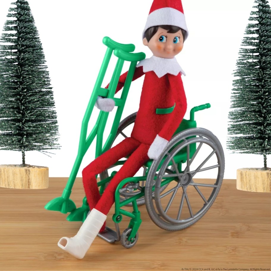 Elf on The Shelf doll in a wheelchair, with crutches, and a cast his foot