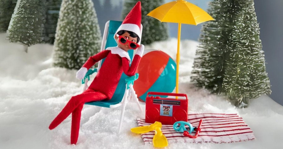 boy Elf on The Shelf doll sitting in a doll size beach chair, beach themed accessories and umbrella around him