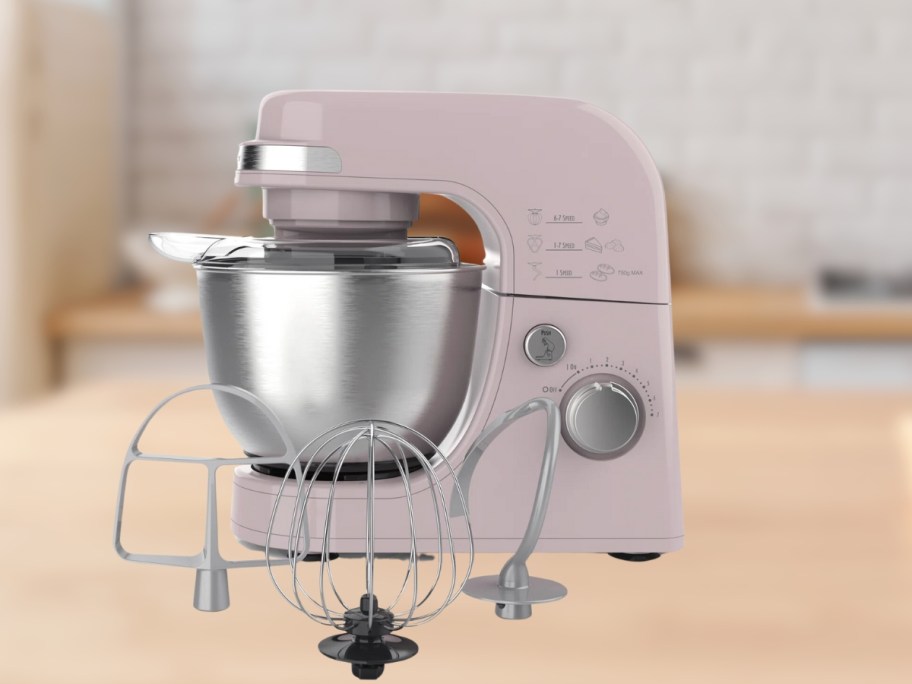 a light pink stand mixer with a stainless bowl and mixer attachments in front of it