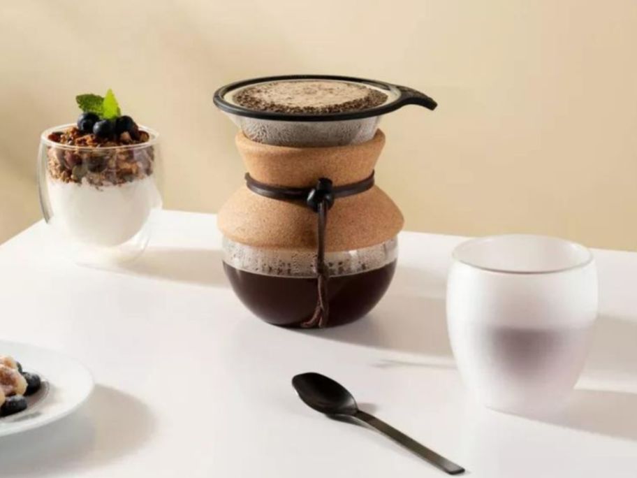 A Bodum pour over coffee maker next to bodum cups and a spoon on a table