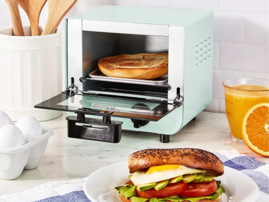 light green Dash Mini Toaster Oven open with bagel inside, sandwich sitting to the side